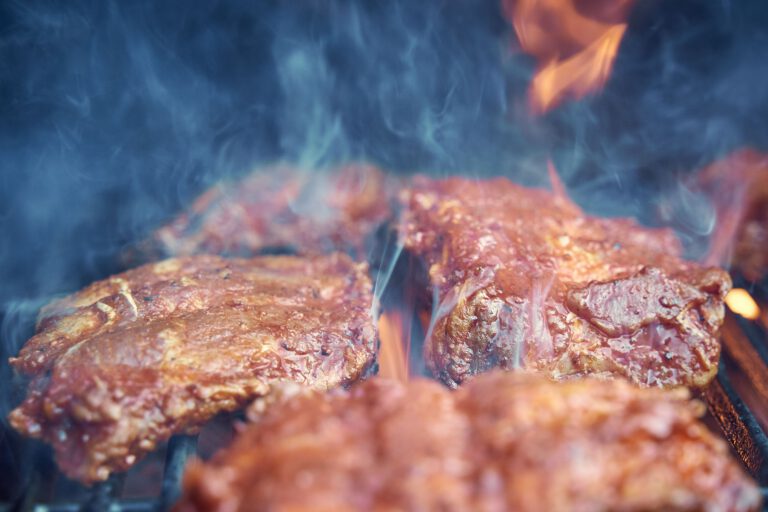 Home Close-up of sizzling raw meat being grilled with smoke and flames on outdoor barbecue.