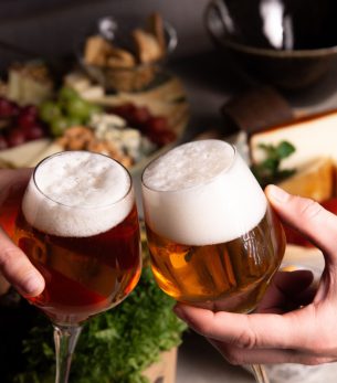 Two people clinking beer glasses over a cheese platter, creating a festive atmosphere.